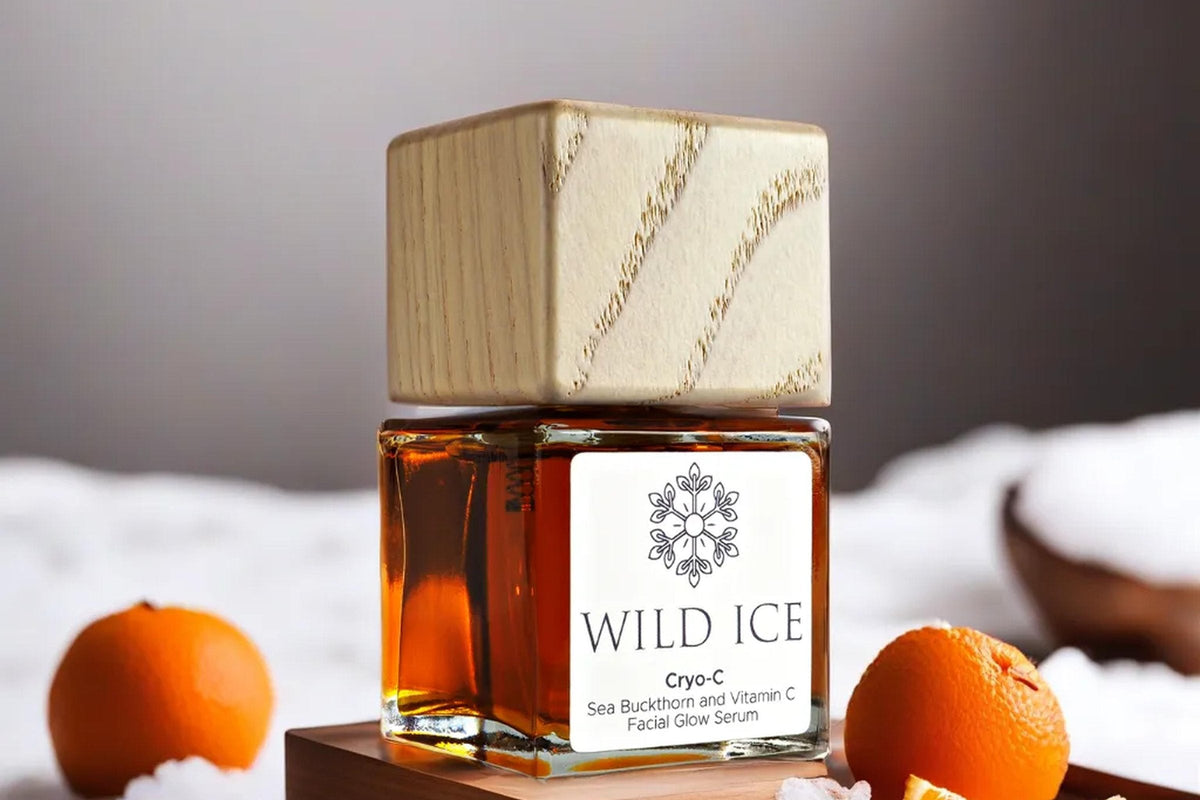 A bottle of Cryo-C sea buckthorn and vitamin C glow serum wearing its wood overcap, standing on a wooden board in snow beside two tangerines and an orange wedge
