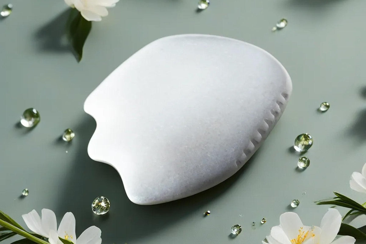 The Mammoth's Tooth white jade gua sha stone seen from a three-quarter angle on a sage-green surface with white blossoms and water drops