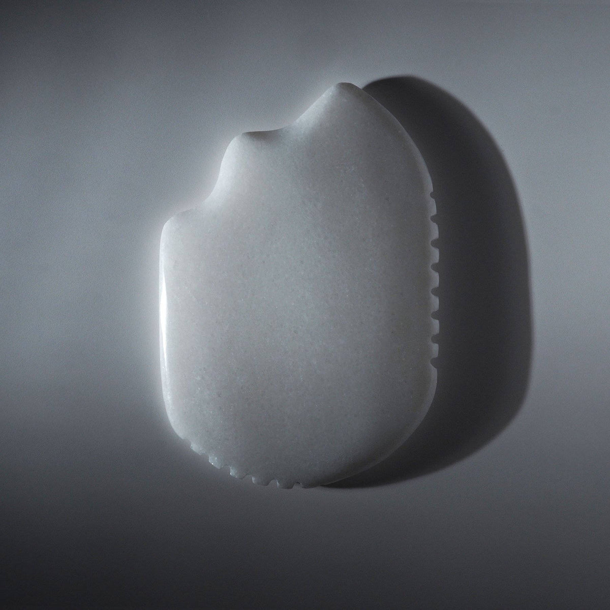 The Mammoth's Tooth white jade gua sha stone lying flat on a pale grey surface, its comb-toothed edge in view