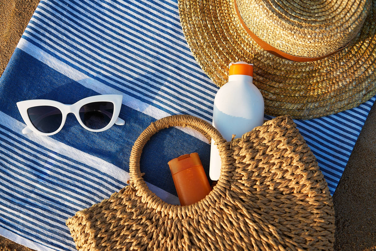A woven straw bag on a striped towel in low golden sunlight with a lotion bottle tucked inside and a white bottle, white sunglasses and a straw hat beside it