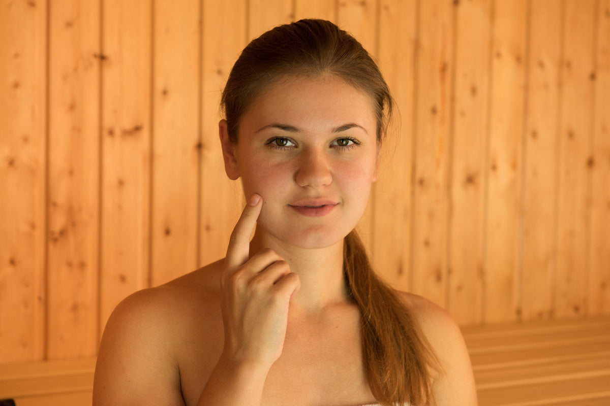 A young woman with flushed cheeks sits in a pine-panelled sauna wrapped in a towel, resting a fingertip on her cheek