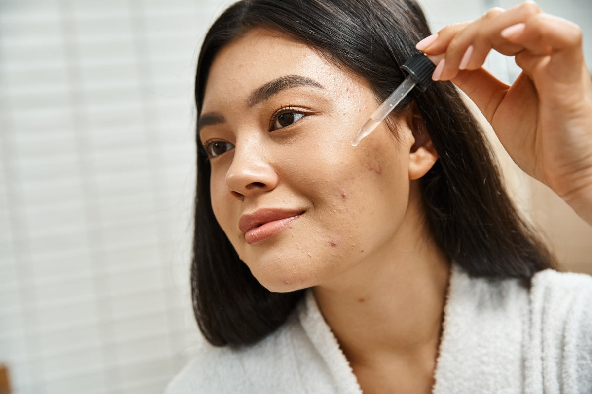 A young woman in a white bathrobe applies a drop of serum from a glass dropper to a cheek with a few small blemishes, in soft bathroom daylight