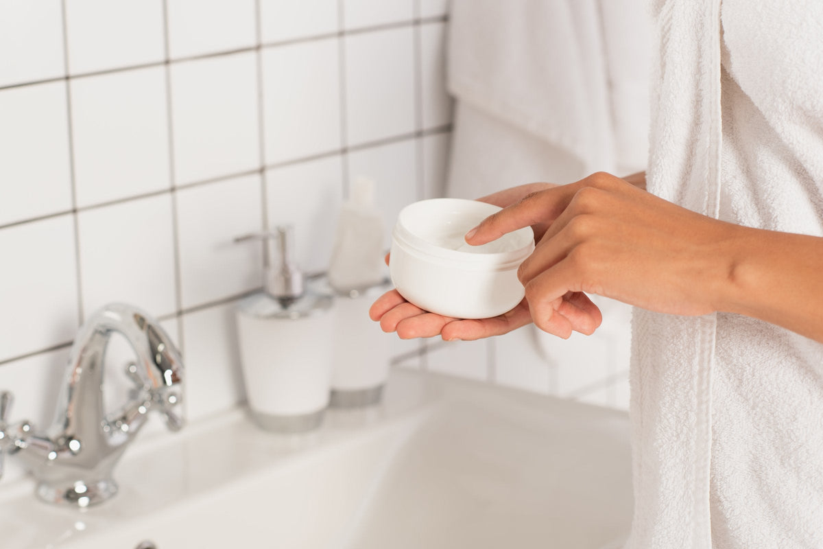 A woman's fingertip touching the cream in an open white jar held over a bathroom sink with a chrome tap and white tiles