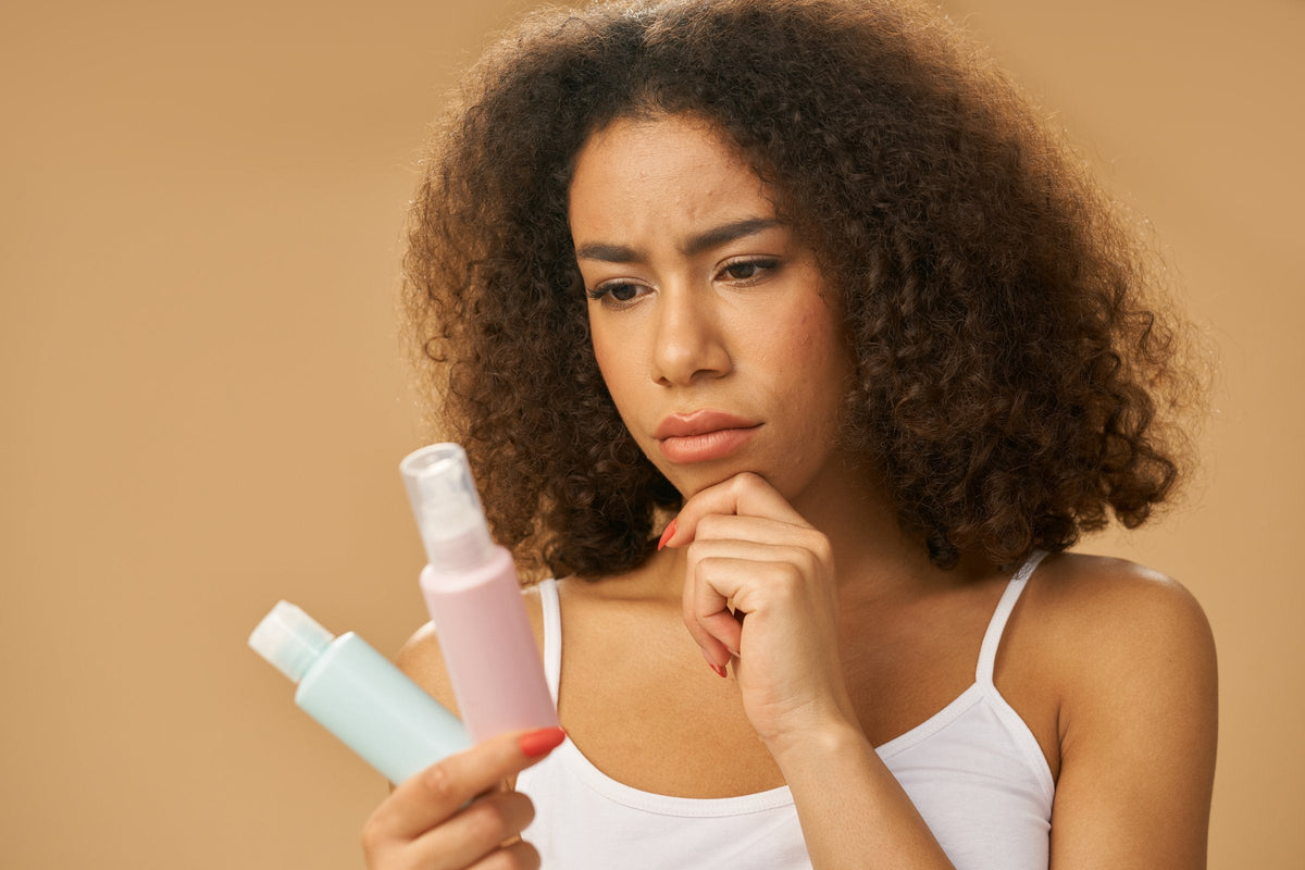 A woman with curly hair rests her chin on her hand and frowns thoughtfully at two small unbranded skincare bottles she is holding, deciding which to use