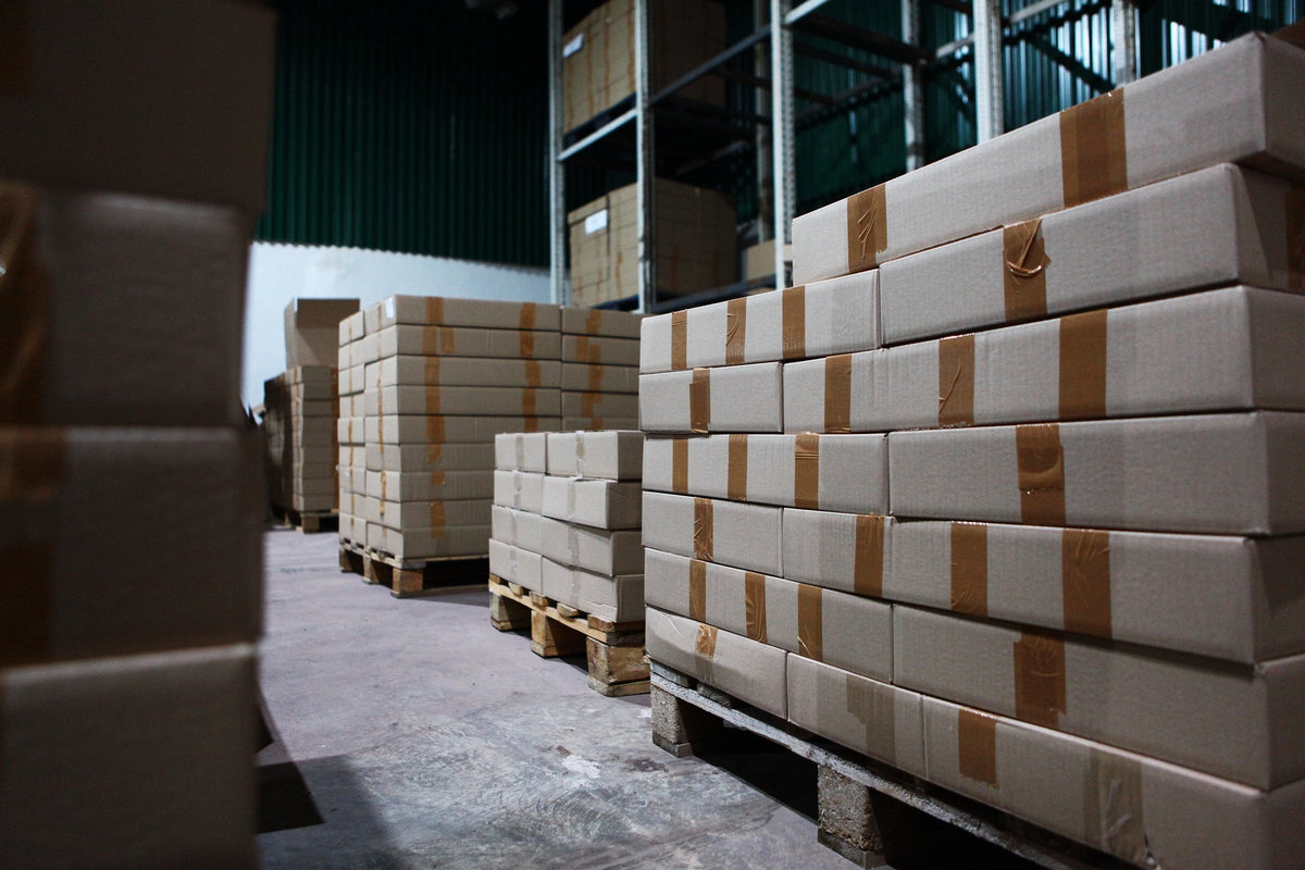 Taped cardboard cartons stacked on wooden pallets along a dim concrete warehouse aisle with steel racking behind