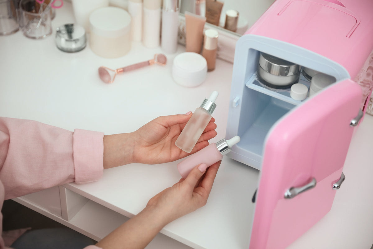 A woman's hands lift a dropper bottle of serum from a small open pink mini fridge on a white vanity beside a mirror and a face roller