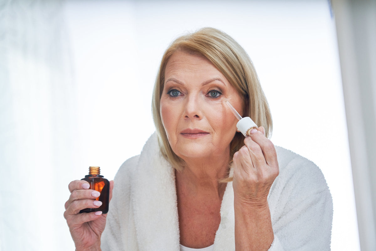A woman in her sixties in a white bathrobe applies a few drops from a glass dropper to her cheek beside a bright bathroom window, the small amber dropper bottle in her other hand