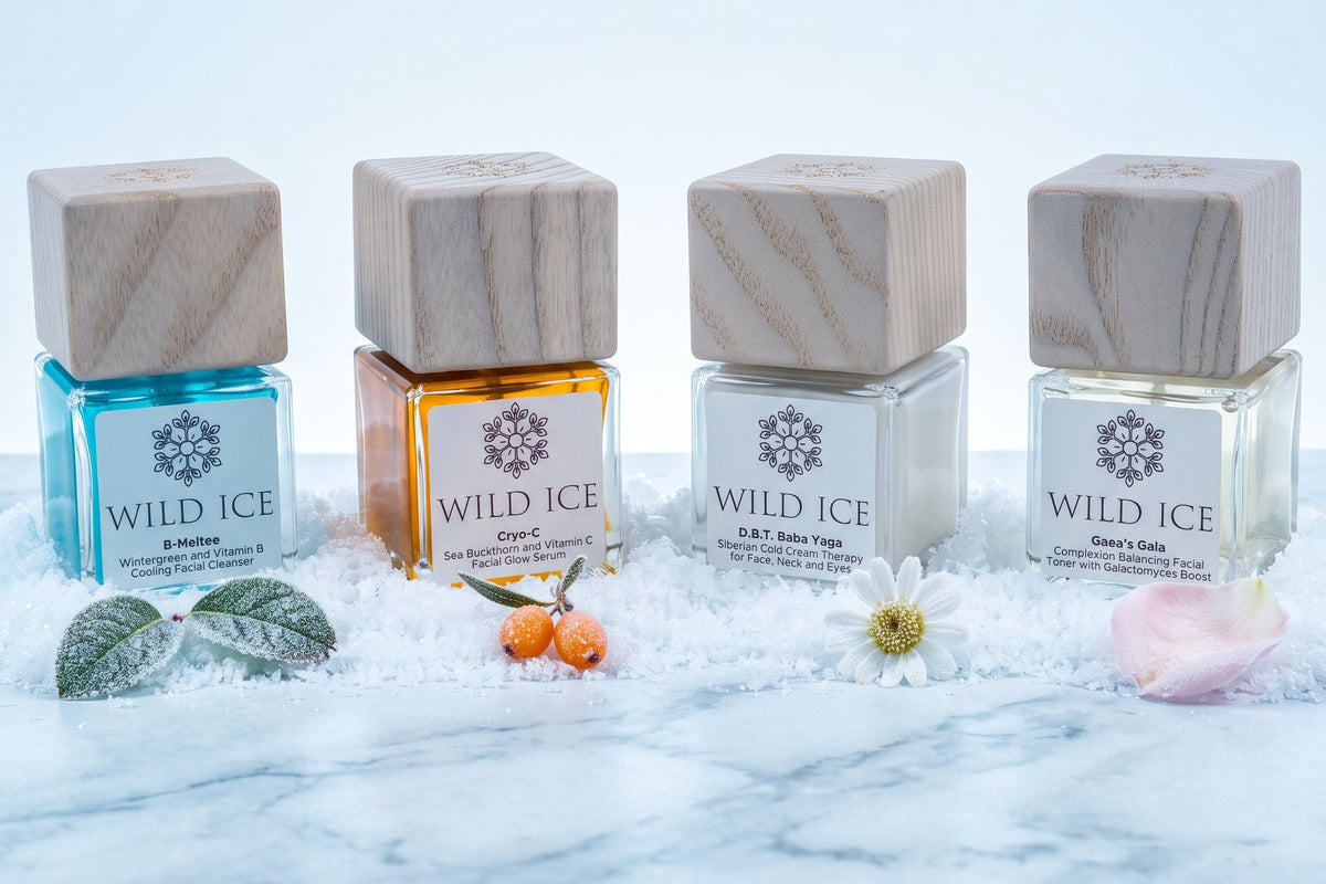 The four full-size Wild Ice Essentials Routine bottles with their wood overcaps on, standing in a row in snow on marble with mint, sea buckthorn berries, a chamomile flower and a rose petal