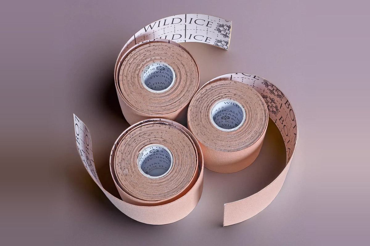 Three rolls of Wild Ice Chrysalis kinesiology face tape in tan on a mauve-grey surface, with strips partly unrolled to show the printed backing