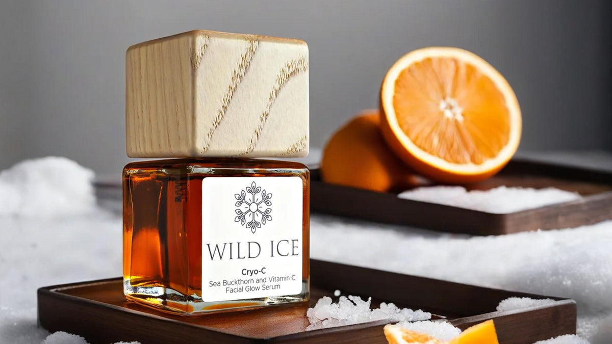 A bottle of Cryo-C sea buckthorn and vitamin C glow serum wearing its wood overcap, on a dark wooden tray in snow, an orange half and a whole orange behind it