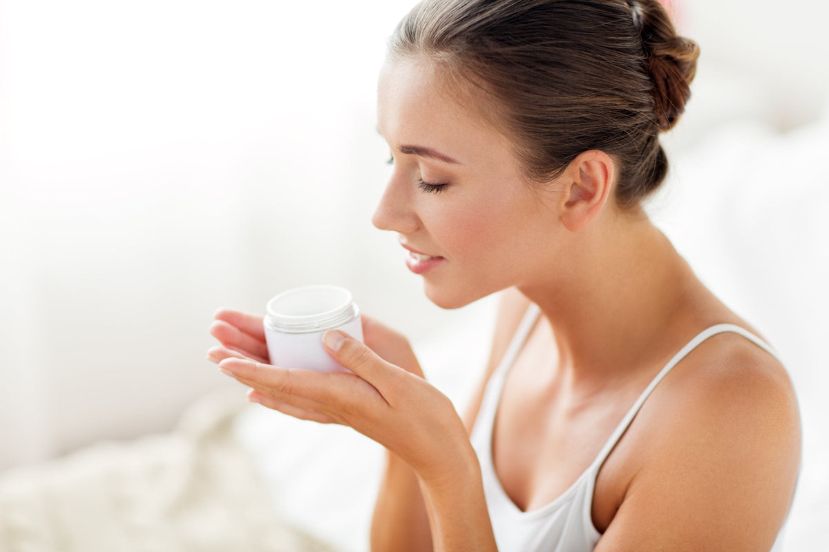 A woman holding an open jar of face cream up to her nose to smell it