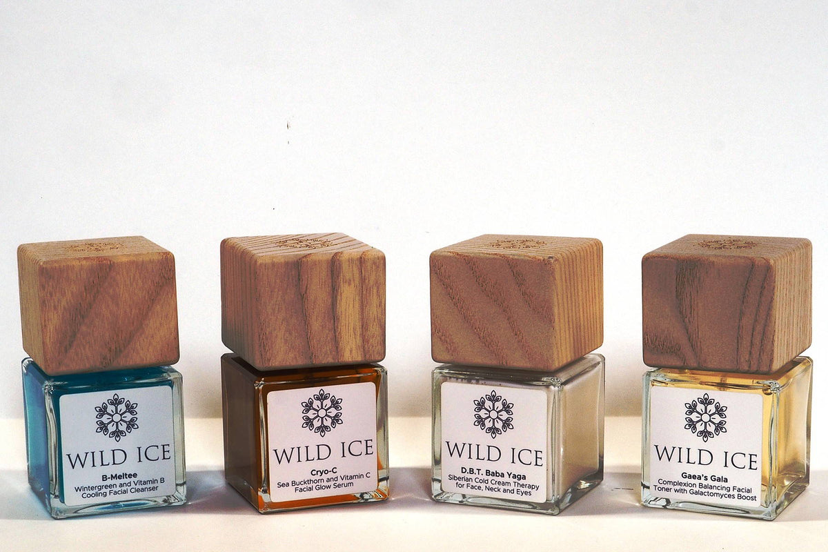 The four full-size Wild Ice Essentials Routine bottles with their wood overcaps on, standing in a row on a plain pale studio surface