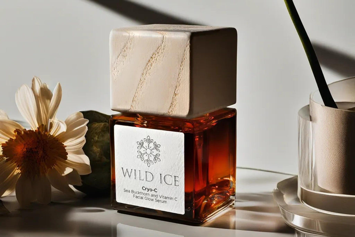 A bottle of Cryo-C sea buckthorn and vitamin C glow serum wearing its pale wood overcap, standing on a reflective surface beside a cream daisy and a glass vase holding a single stem, with long shadows