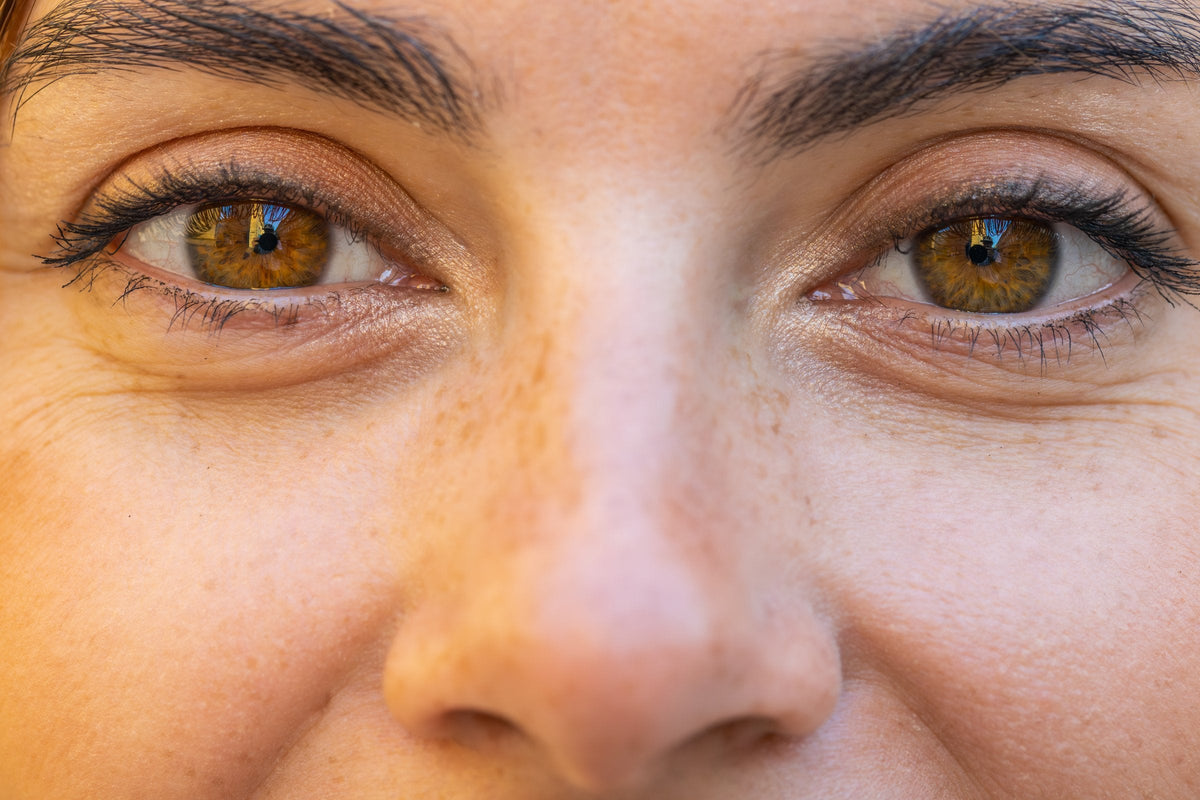 A close view of a woman's brown eyes as she smiles softly, with faint fine lines beneath them and natural freckled skin in daylight