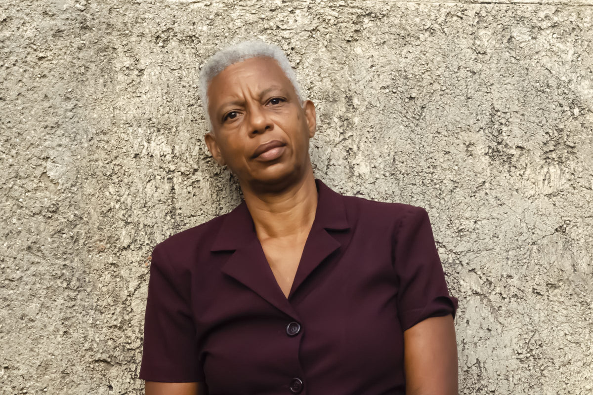 A calm older woman with cropped white hair, natural fine lines and wrinkles, standing against a rough plaster wall in daylight