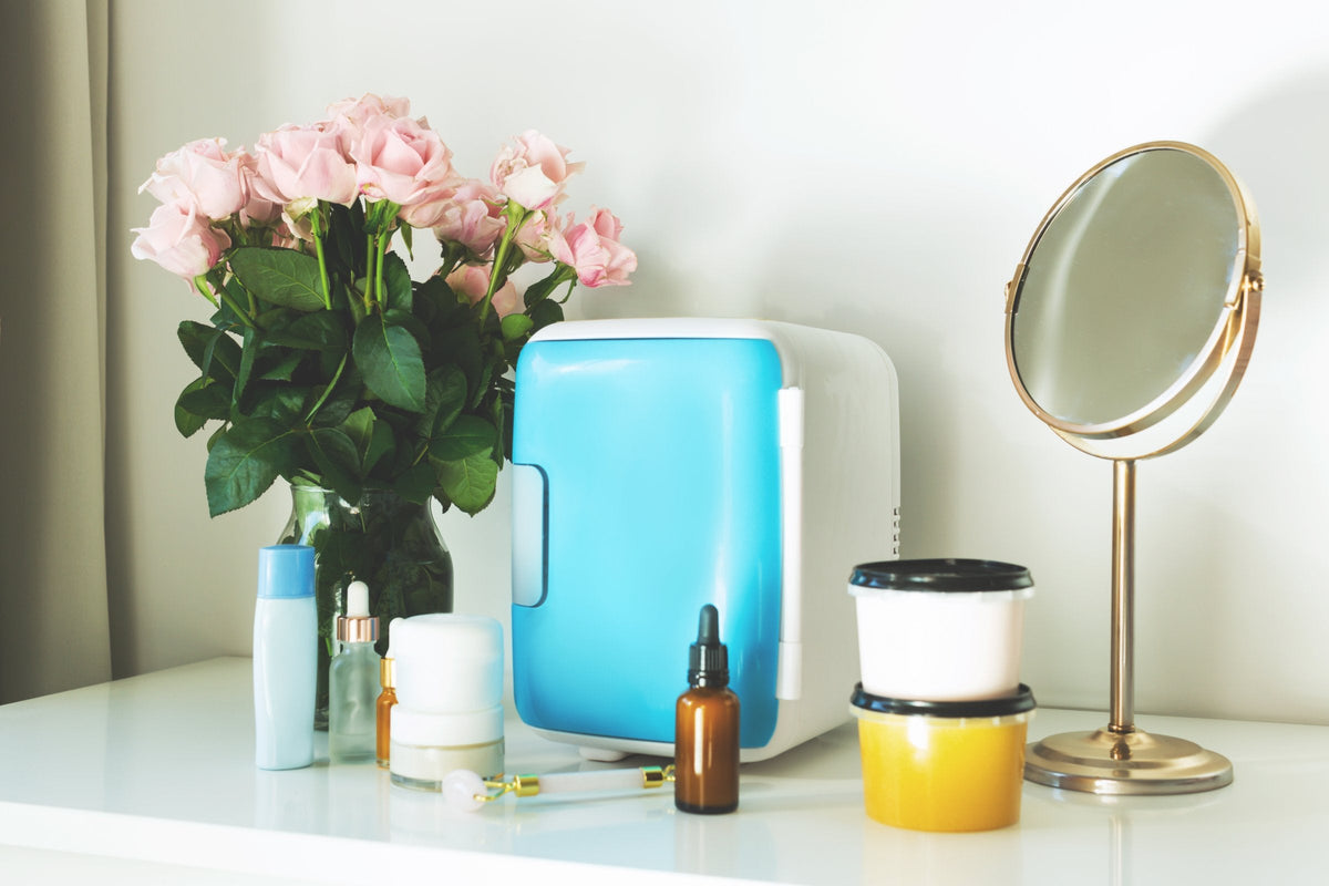 A small mini fridge on a vanity table with skincare bottles, a mirror and fresh flowers