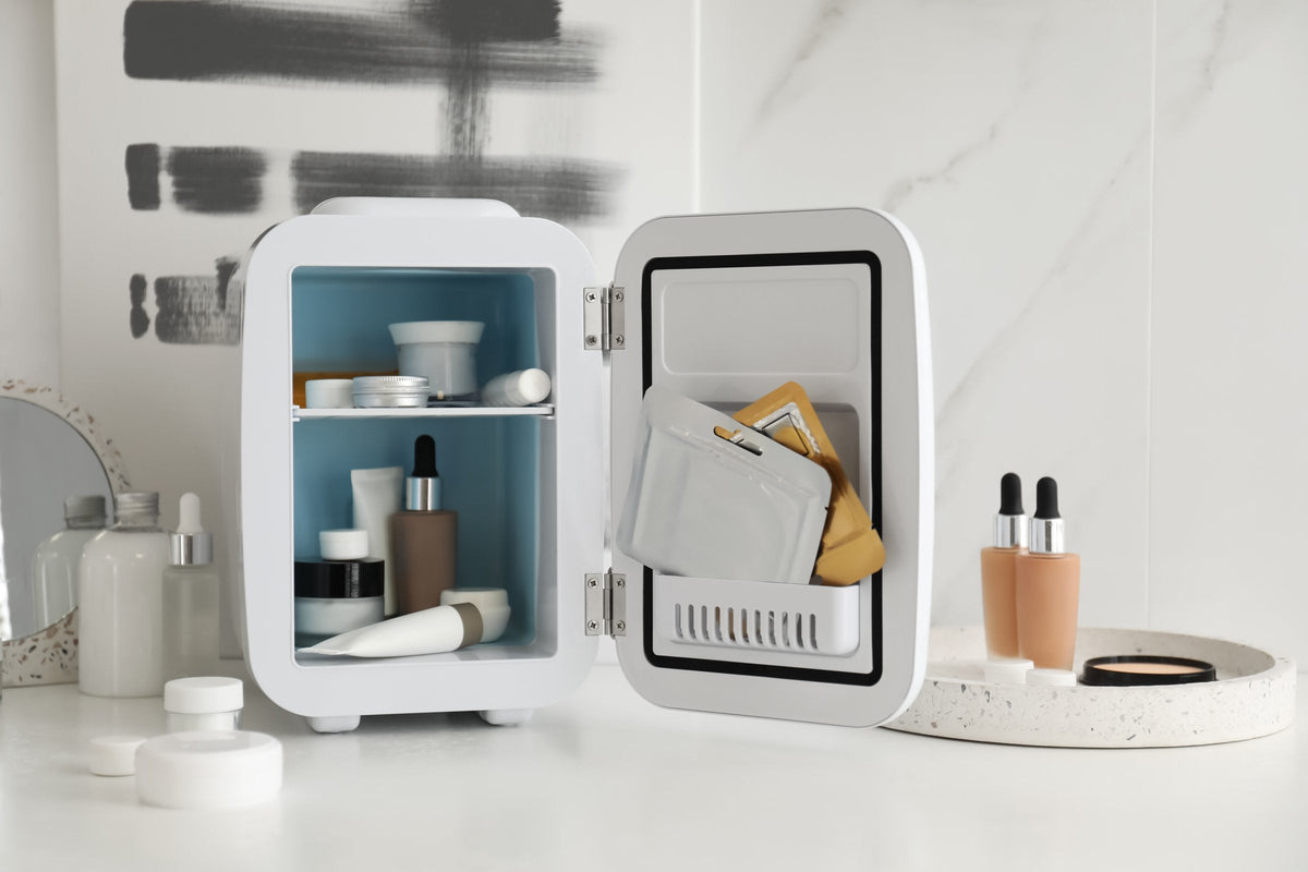 A small white skincare fridge stands open on a bathroom counter with cream jars and a serum bottle on its shelves and two dropper bottles waiting beside it