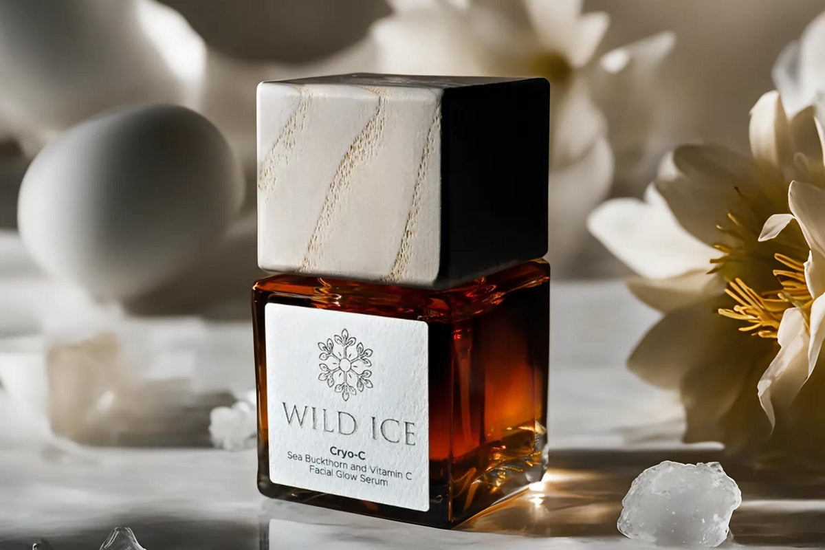 A bottle of Cryo-C sea buckthorn and vitamin C glow serum wearing its wood overcap, standing on marble among white flowers and chips of ice