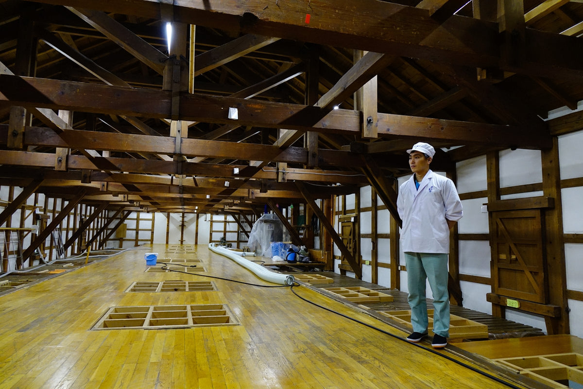 A brewer in a white work coat and cap stands in the timber-beamed hall of a sake brewery in Akita, Japan, beside rows of square hatches set into the wooden floor
