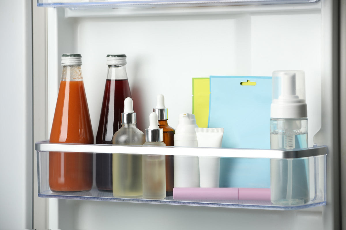 Two dropper serum bottles and small cream jars chill in a refrigerator door rack next to juice bottles and a couple of sheet mask sachets