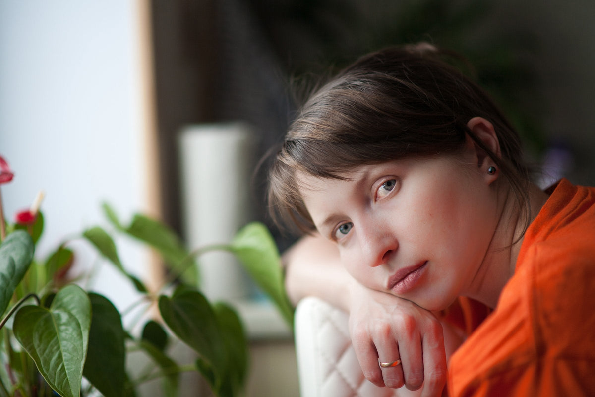 A woman with a tired, makeup-free complexion resting her chin on her hand at home, looking at the camera in soft window light beside a houseplant