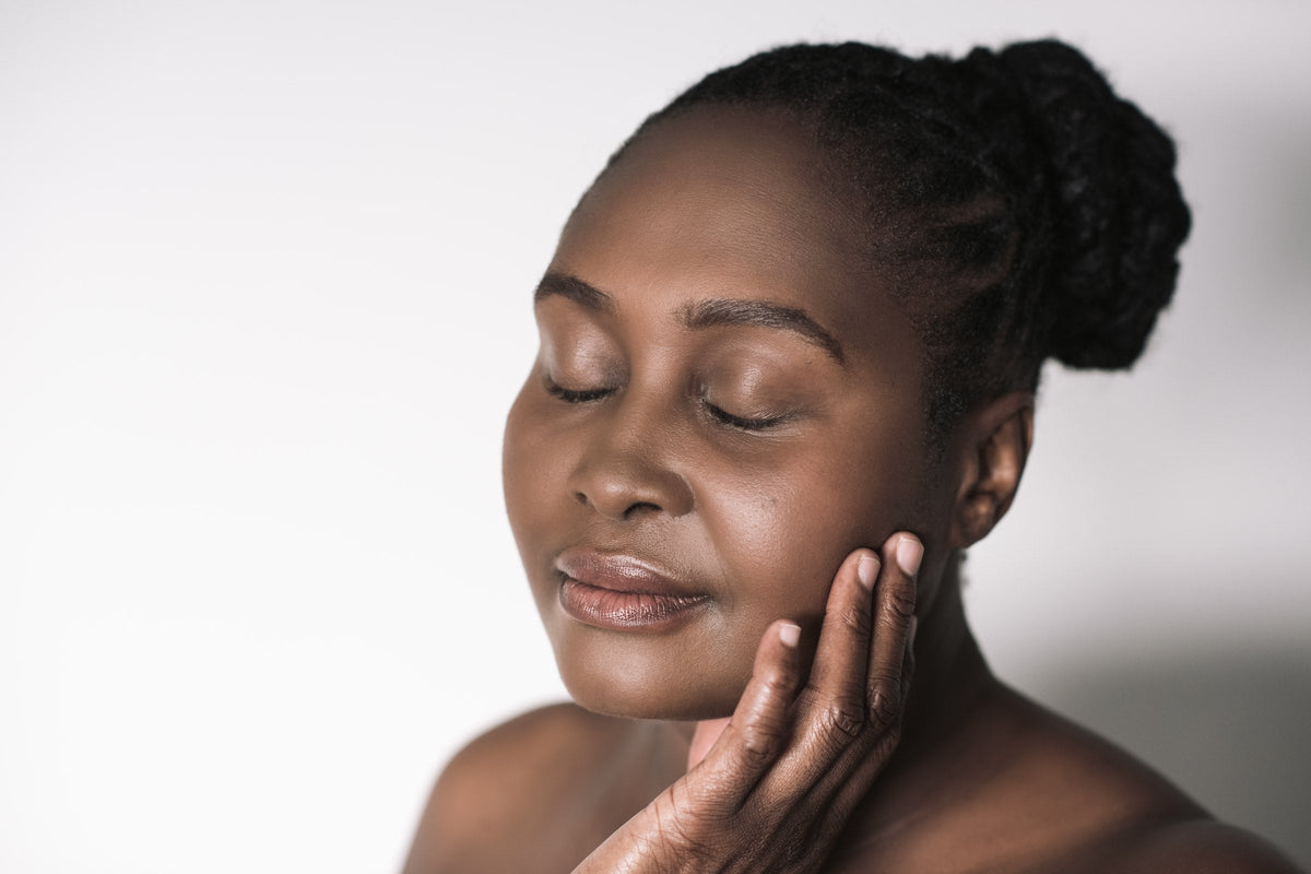 A woman with her eyes closed rests the fingertips of one hand against her cheek, photographed against a plain white background