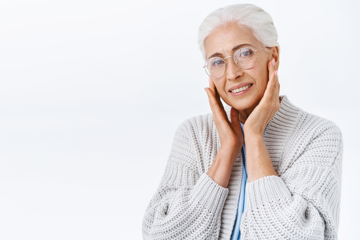 A smiling older woman with grey hair and glasses, hands resting on her cheeks, against a white background