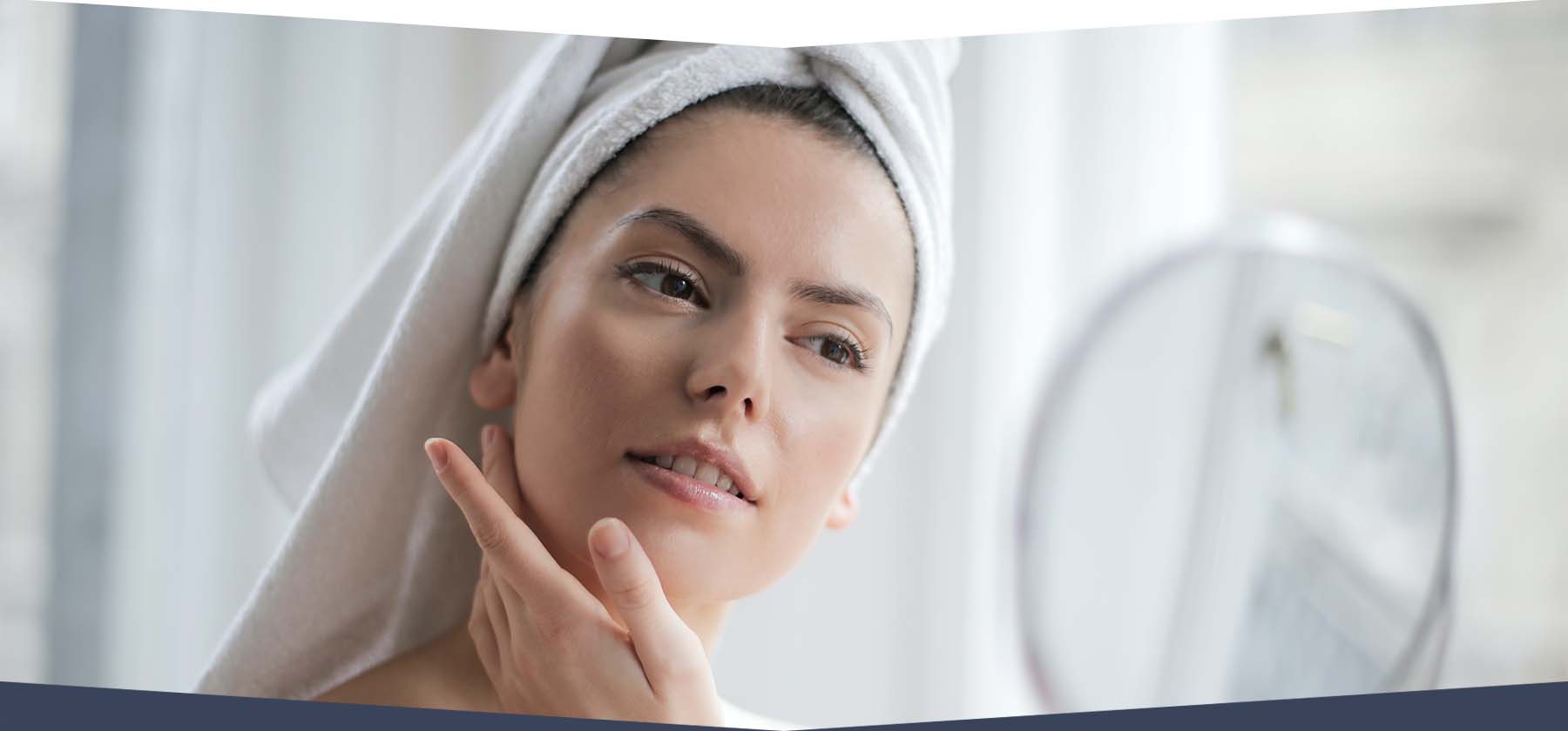 Woman with hair wrapped in white towel looking into mirror touching skin