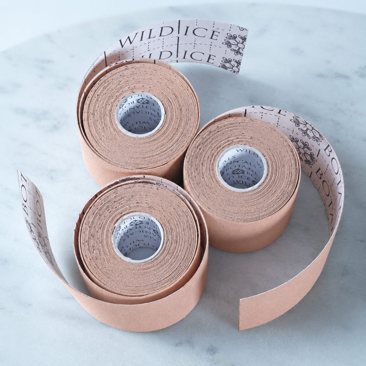 Three rolls of beige cotton Chrysalis kinesiology face tape, partially unrolled on marble with printed cut guide lines