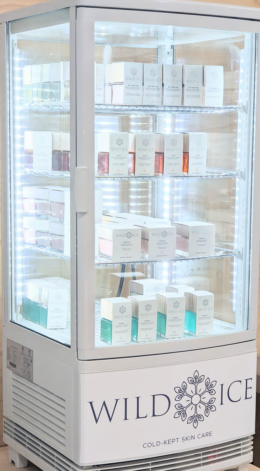 A lit glass display cooler stocked with boxed Wild Ice products on four shelves, with WILD ICE COLD-KEPT SKIN CARE printed across its base